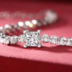 Princess-cut diamond tennis bracelet with round accent stones, displayed on a soft red background