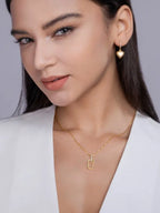 Woman wearing gold jewelry including a necklace and heart-shaped earrings against a neutral background