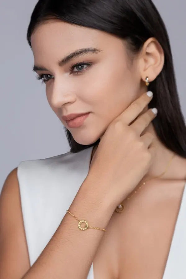 Woman wearing gold jewelry including earrings and a bracelet on a plain background