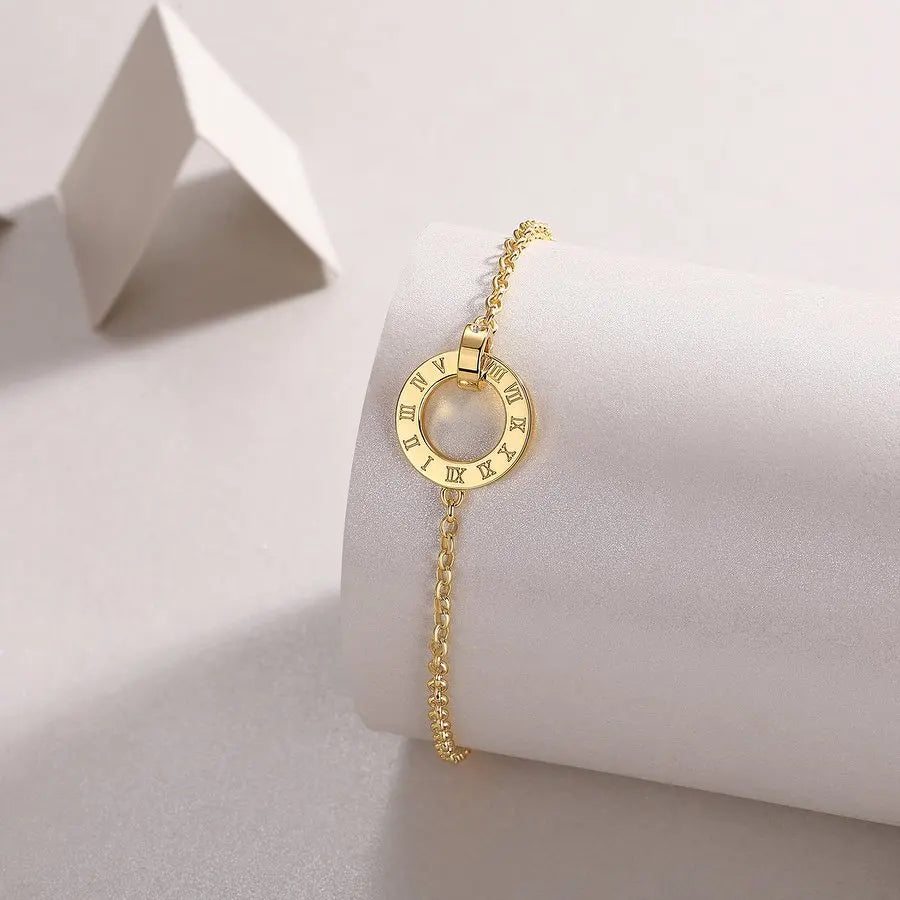 Yellow gold bracelet with a circular Roman numeral charm displayed on a minimalist white jewelry stand