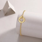 Yellow gold bracelet with a circular Roman numeral charm displayed on a minimalist white jewelry stand