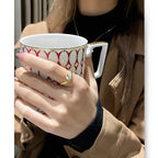 Hand holding a patterned mug with a gold ring, wearing a beige coat.